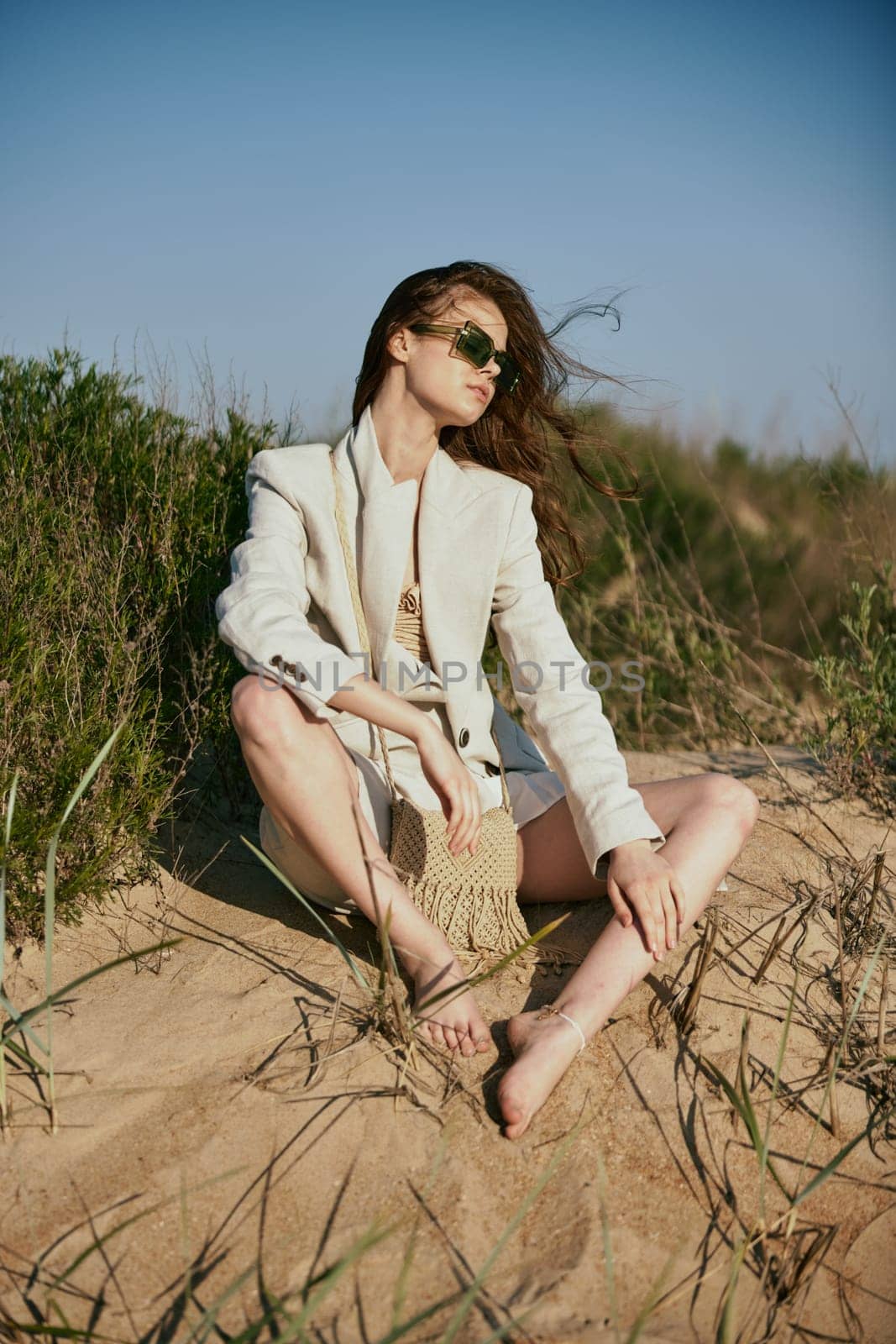 a beautiful woman in a light, fashionable jacket, in sunglasses sits barefoot on the sand in windy weather. High quality photo