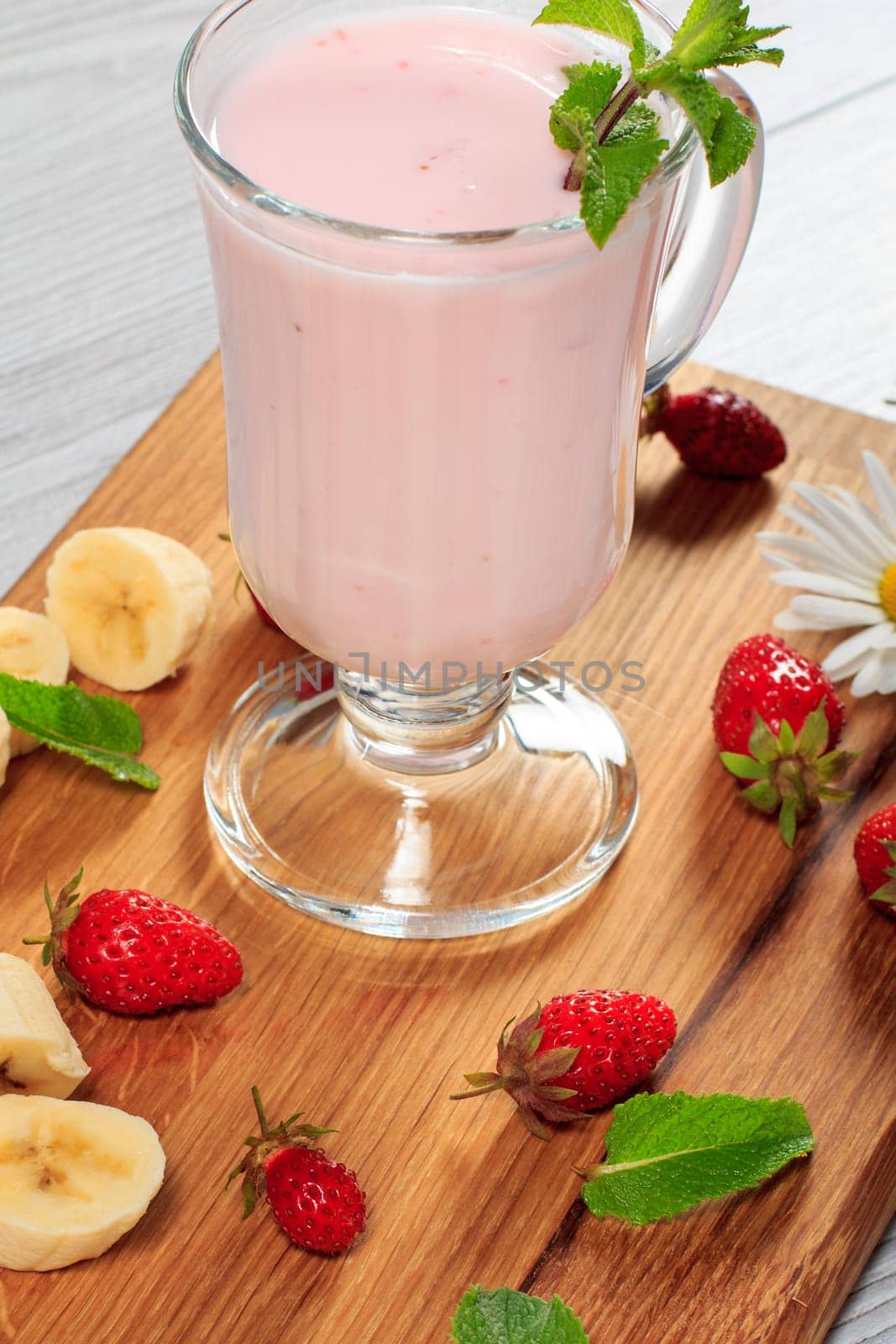 Glass of delicious milk shake with mint and fresh strawberries, banana, chamomile on wood cutting board