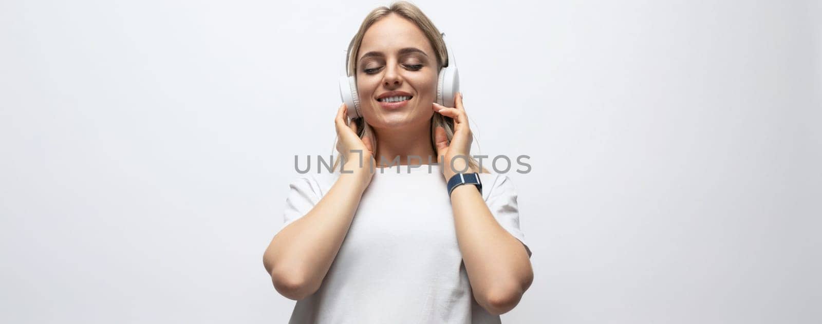 horizontal photo of a young woman in a white tank top listening to a podcast in wireless headphones on a white background with free space.