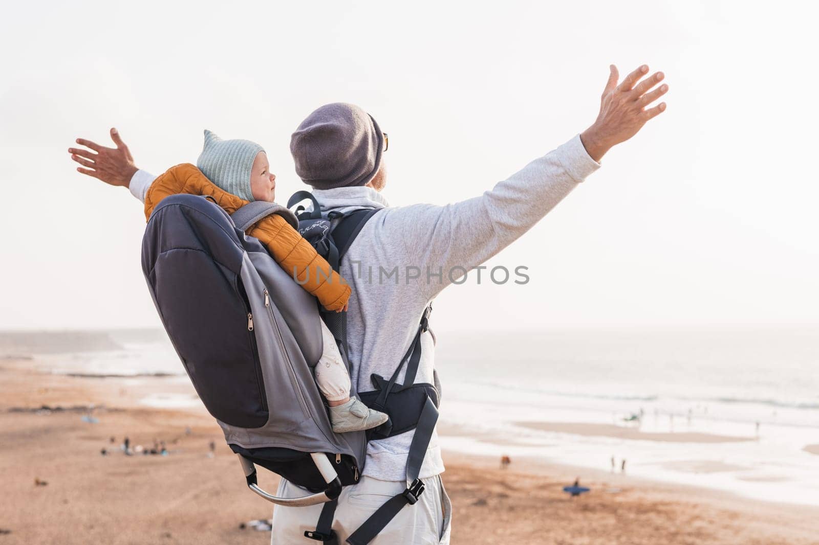 Young father rising hands to the sky while enjoying pure nature carrying his infant baby boy son in backpack on windy sandy beach. Family travel concept. by kasto