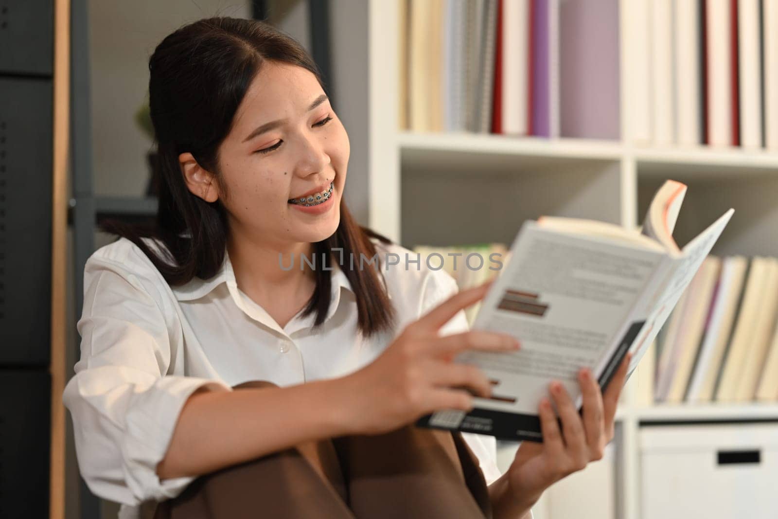 Cheerful Asian woman in casual outfit reading book in cozy living room, enjoying leisure time on weekend at home by prathanchorruangsak