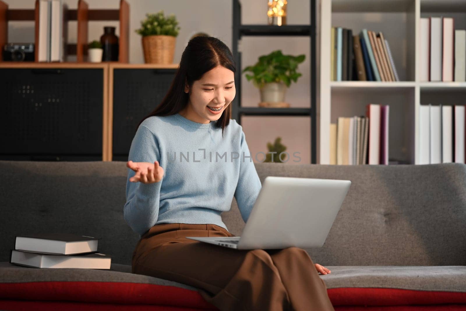 Cheerful young woman sitting on couch at home, using laptop for distant studying, online communication.