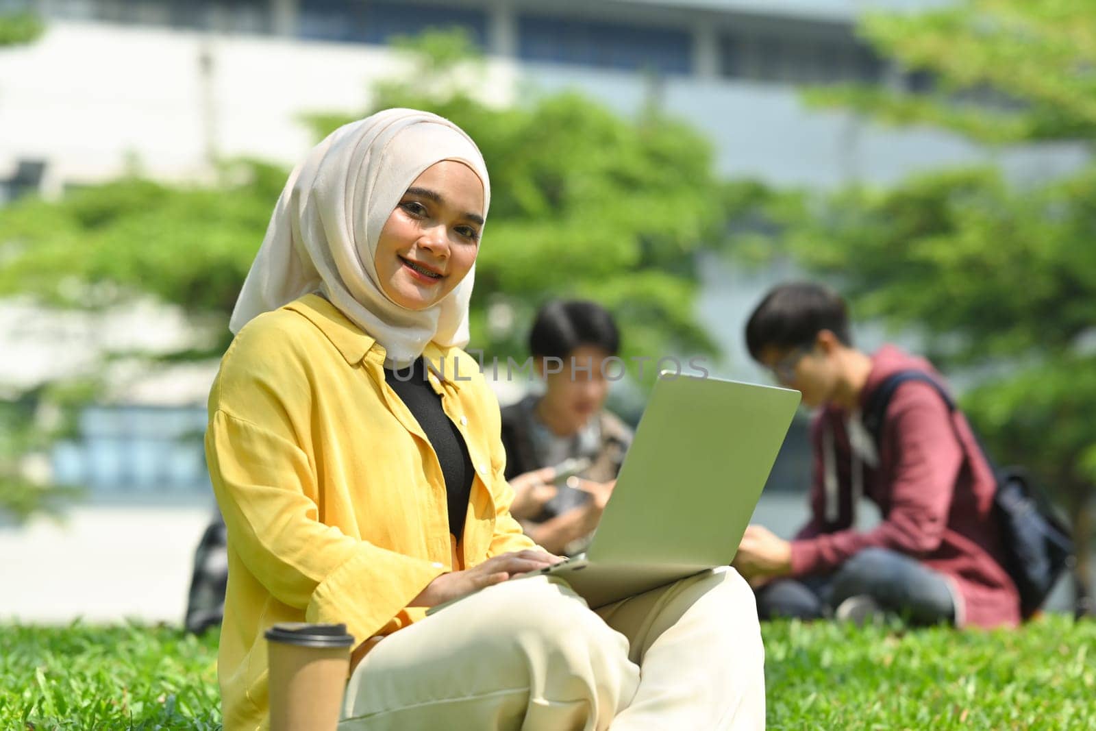 Smiling muslim college student using laptop on green grass in the campus. Education, technology and lifestyle concept.