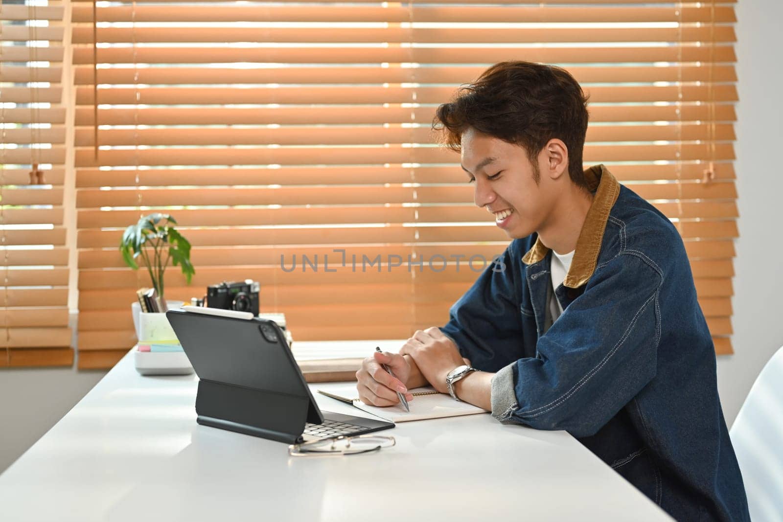 Smiling asian man student looking at digital tablet screen and making notes on notebook. Work from home, e-learning concept by prathanchorruangsak