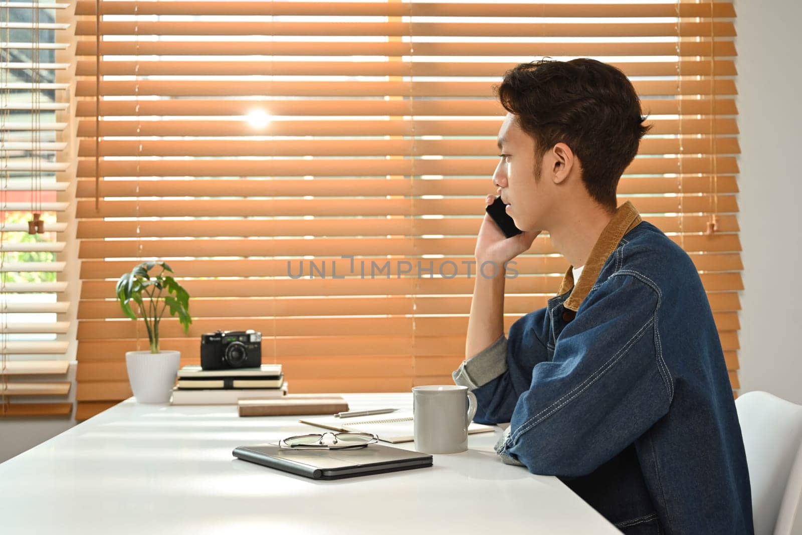 Side view of young asian male freelancer sitting in modern home office interior and talking on mobile phone.