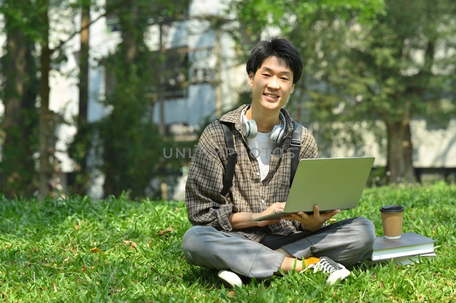 Smiling asian man student using laptop on green grass in front of university building. Education, technology and lifestyle concept.