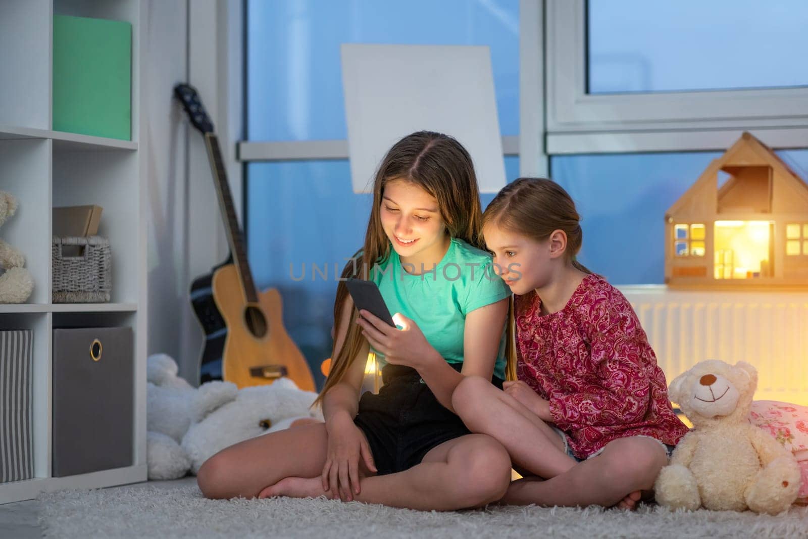 Happy sisters playing with smartphone sitting on floor in kids room in evening