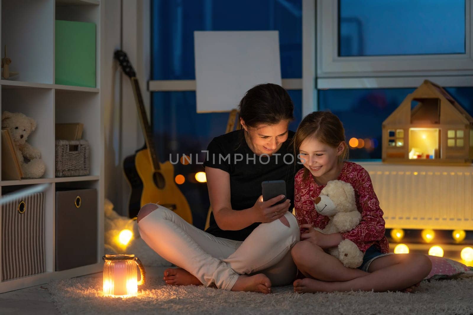 Smiling mother with little daughter using smartphone sitting on floor in illuminated night room