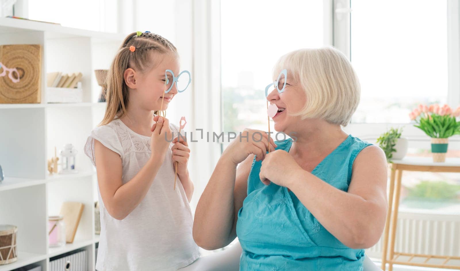 Cute granddaughter and granny holding glasses and lips on sticks in bright room