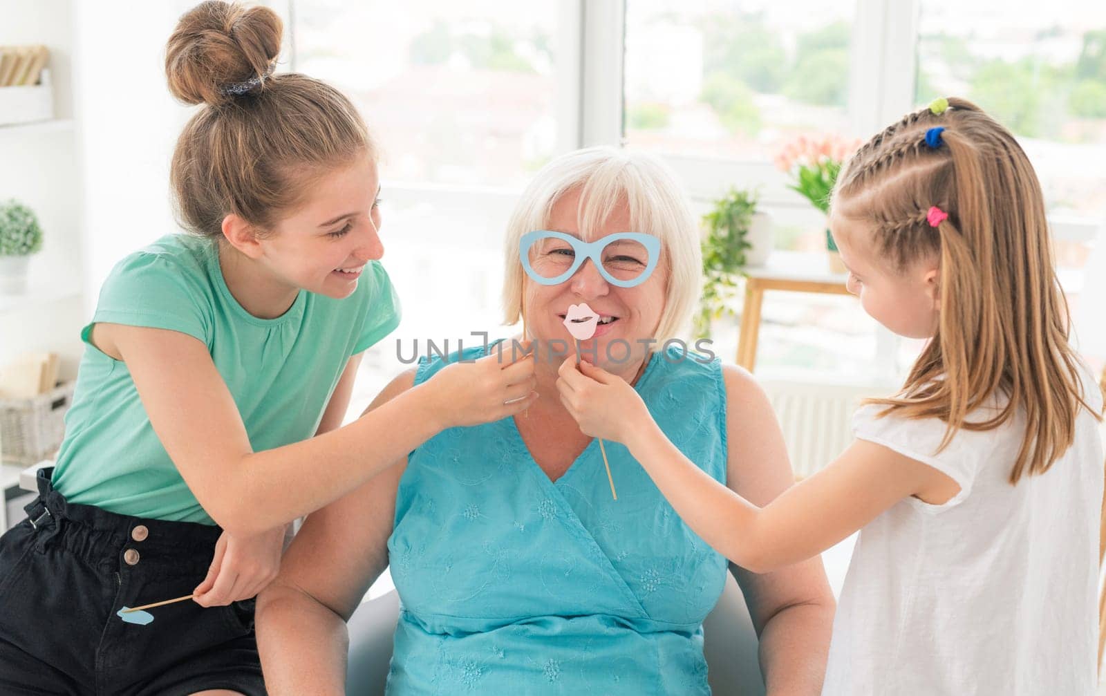 Cute granddaughters putting paper glasses and lips on smiling grandmother at home