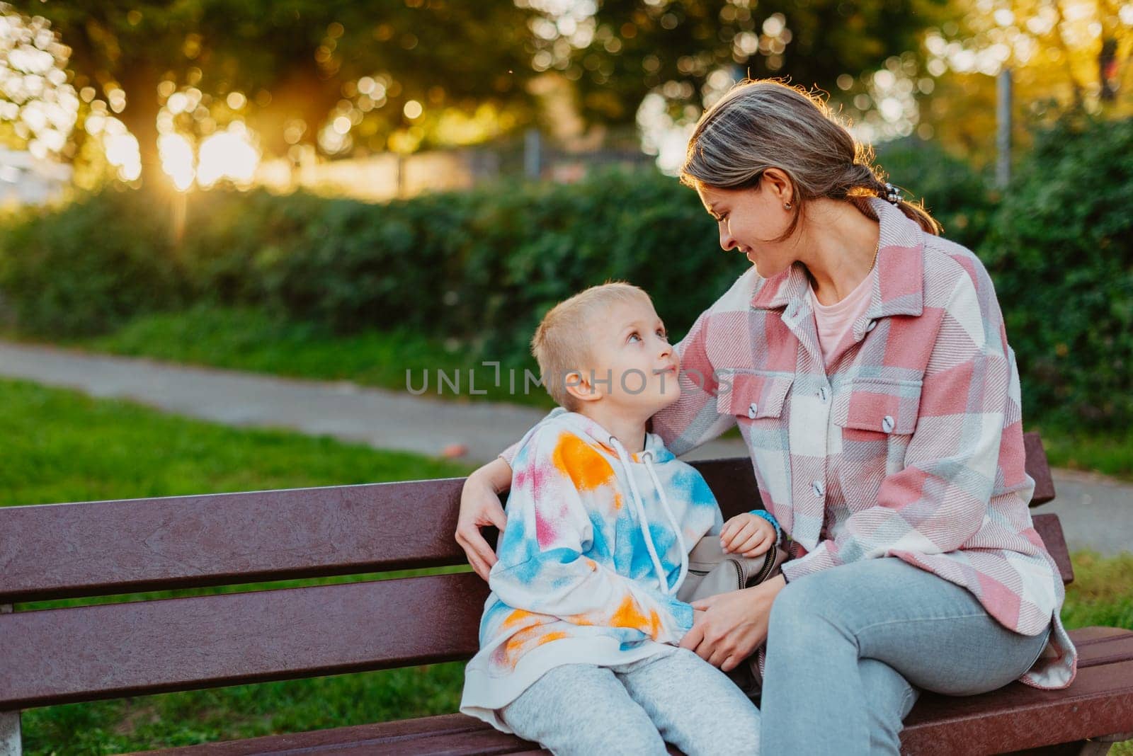 mother and son sit on a park bench in the rays of the setting sun. the concept of a family. Mother's Day. beautiful girl (mother) with a boy (son) in the park in the park are sitting on a bench at sunset.