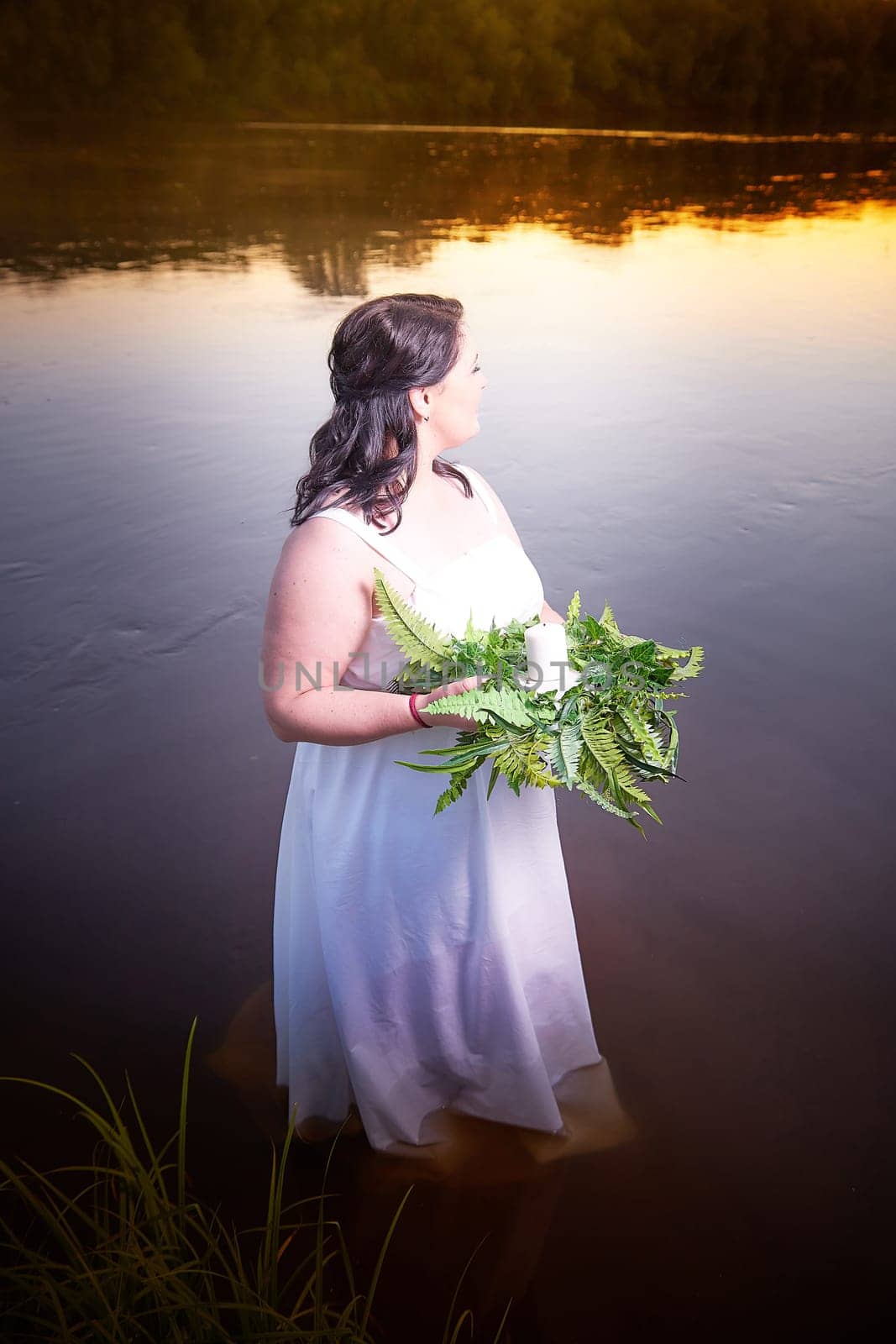 Slavic plump plump chubby girl in long white dress on the feast of Ivan Kupala with flowers and water in a river or lake on a summer evening by keleny