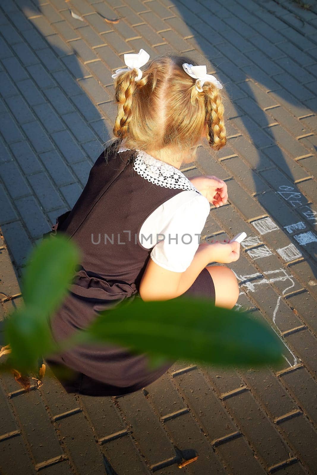 Little girl of elementary school student in modern school uniform drawing with chalk on asphalt outdoors. Female child schoolgirl going to school. Back to school in september 1 in Russia by keleny