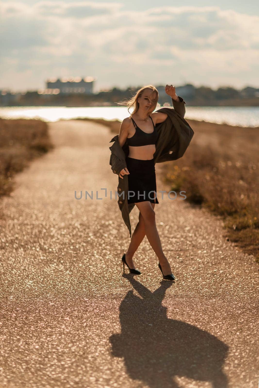 Portrait blonde sea cape. A calm young blonde in an unbuttoned khaki raincoat walks along the seashore, under a raincoat a black skirt and top by Matiunina