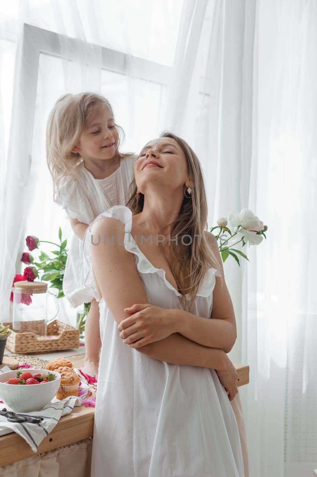 A little blonde girl with her mom on a kitchen countertop decorated with peonies. The concept of the relationship between mother and daughter. Spring atmosphere