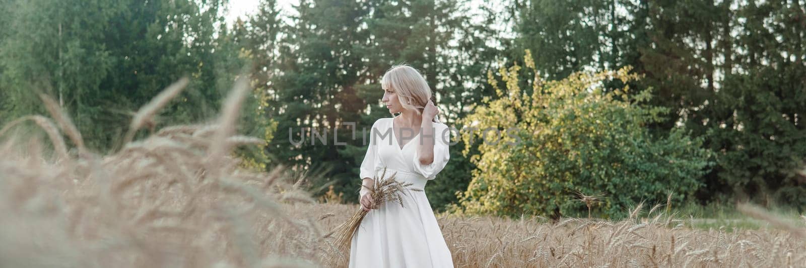 A blonde woman in a long white dress walks in a wheat field. The concept of a wedding and walking in nature.