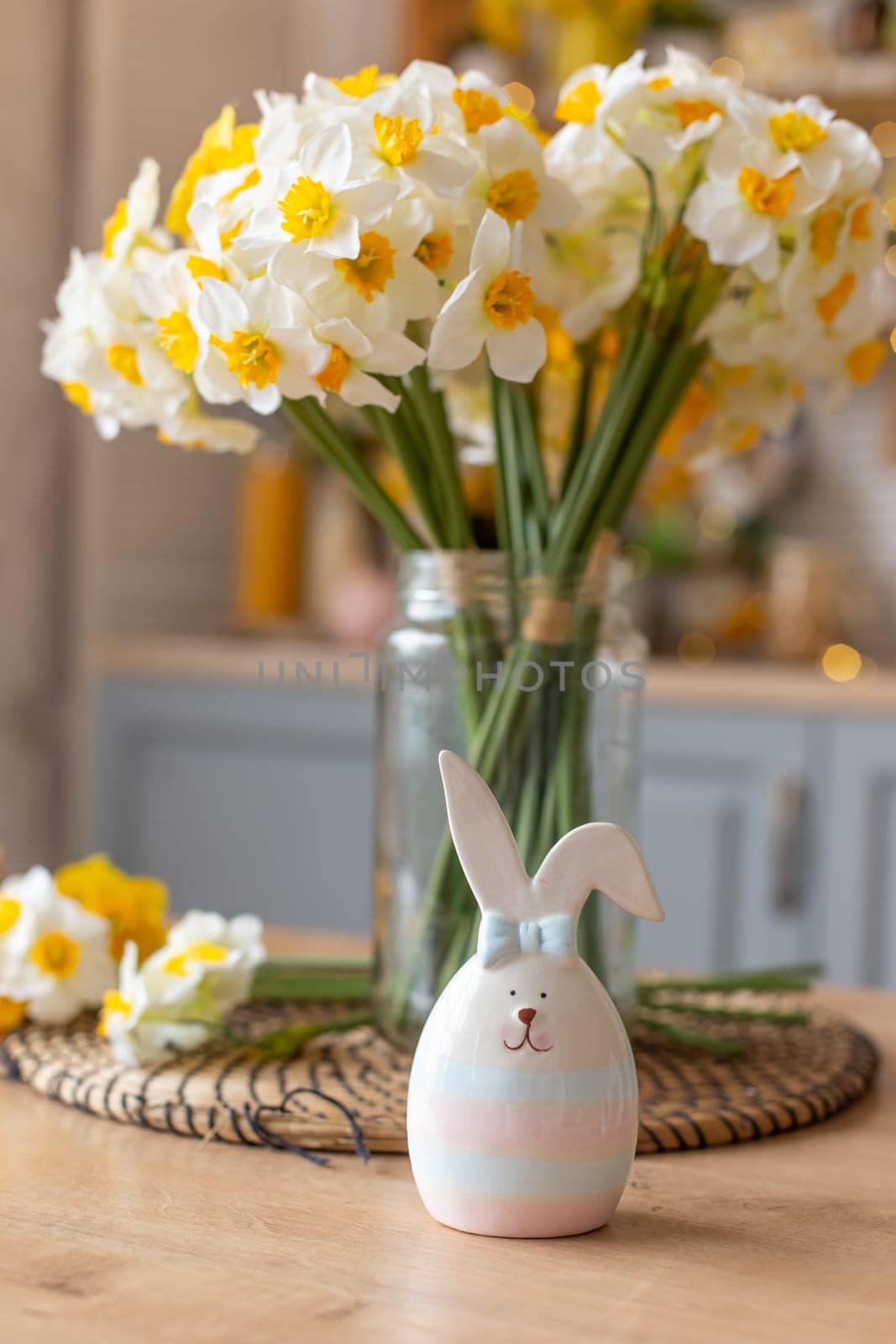 A ceramic figurine of a cute striped hare stands on the kitchen table next to a glass vase with daffodil flowers. Close up. Vertical