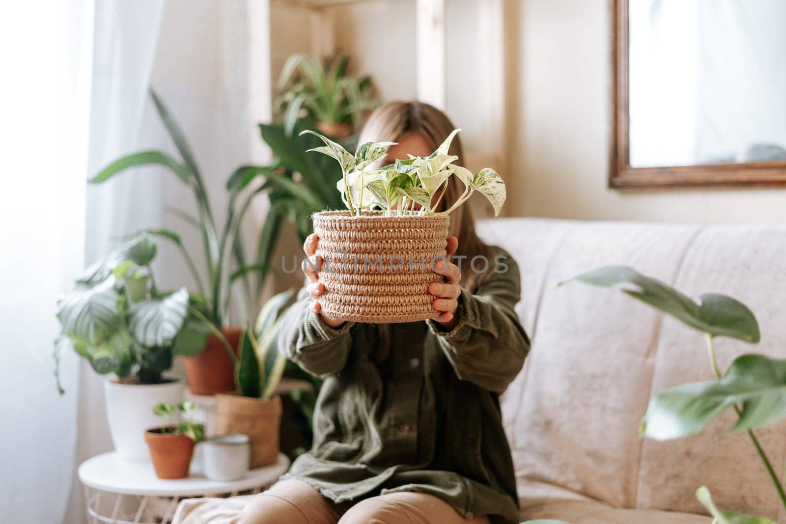 Woman holding wicker basket with plant golden pothos epipremnum, hiding face, sitting and relaxing on the couch with a lot of plants. Cozy home and urban jungle concept