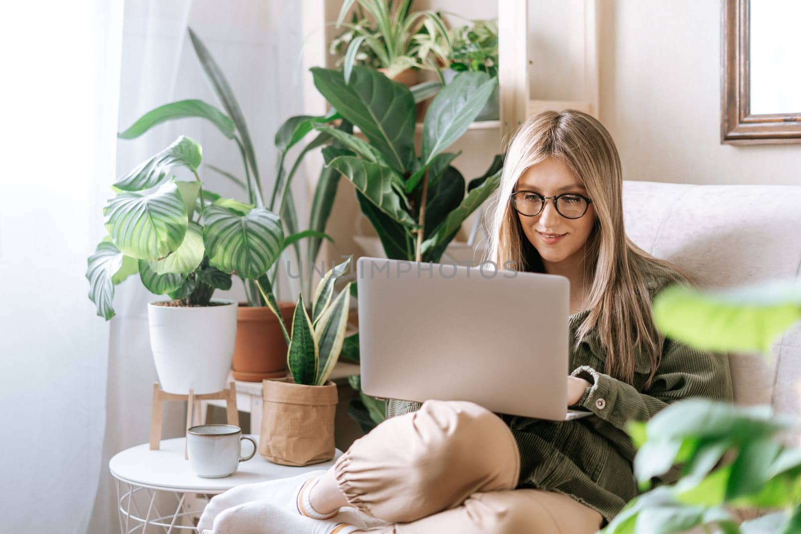 Freelance woman in glasses typing at laptop and working from home office. Happy girl sitting on couch in living room with plants in cozy urban jungle home. Distance learning online education and work by Ostanina