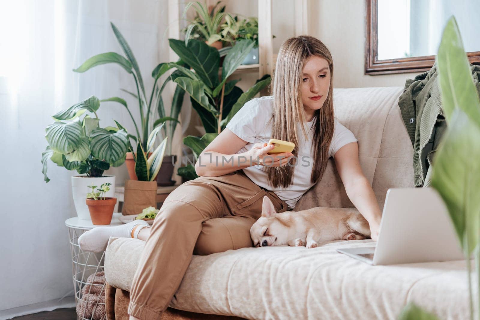 Freelance woman with sleeping dog and laptop, texting at mobile phone and working from home office. Happy girl sitting on couch in living room with plants. Distance learning online education and work by Ostanina