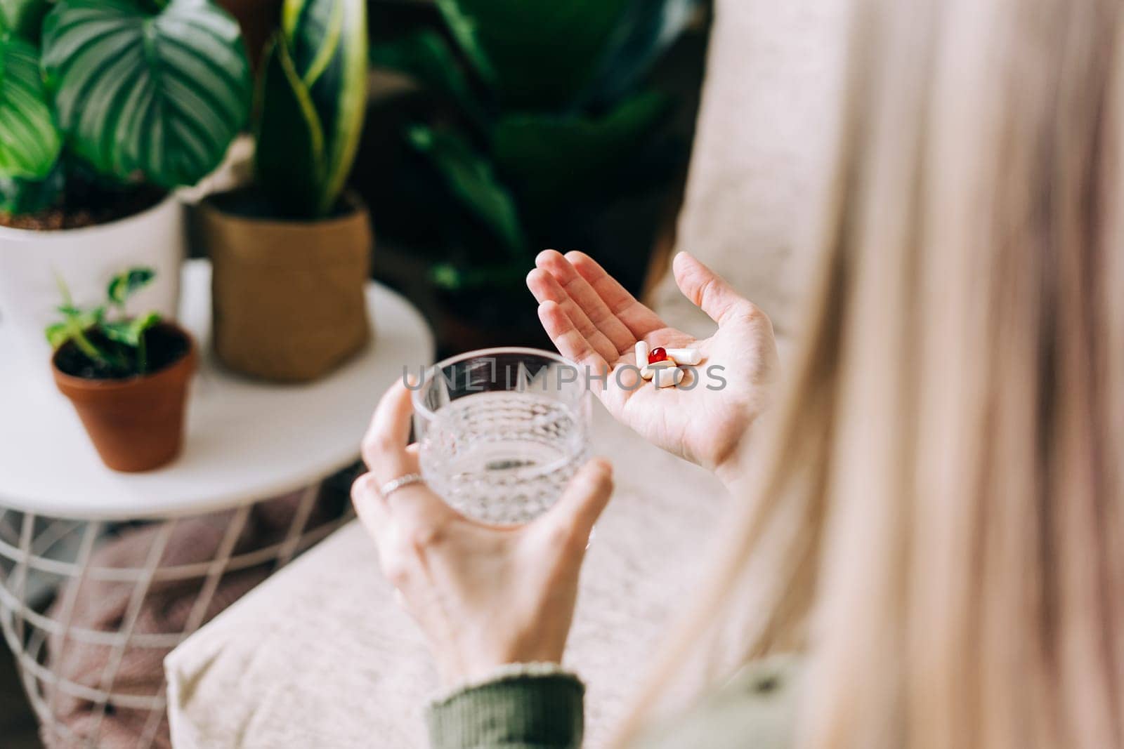 Close up of woman hands holding glass of water and tablet vitamin pills. Girl in living room with plants at home, healthy organic lifestyle concept.