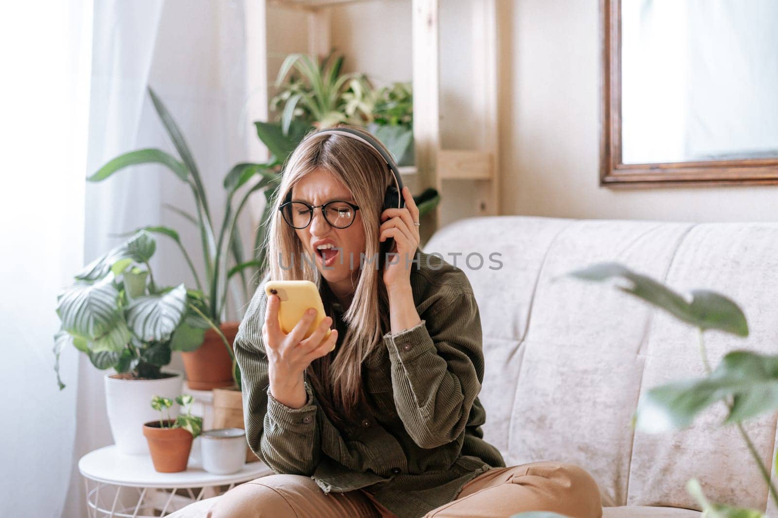 Freelance woman in glasses with mobile phone listening music in headphones and relax at home. Happy girl sitting on couch in living room with plants. Urban jungle concept