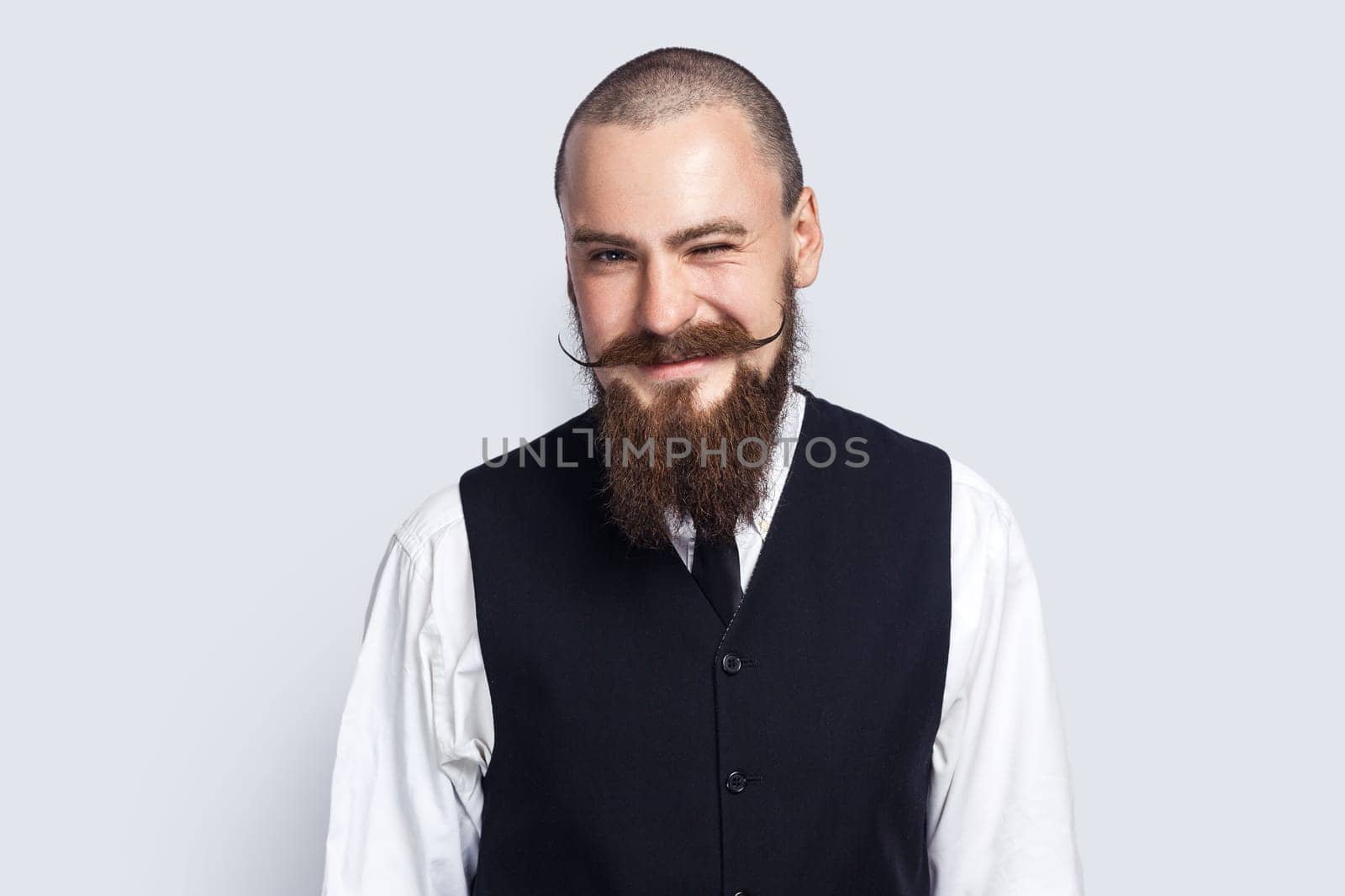 Portrait of positive happy handsome man with beard and mustache standing and winking to camera, flirting with somebody, smiling happily. Indoor studio shot isolated on gray background.