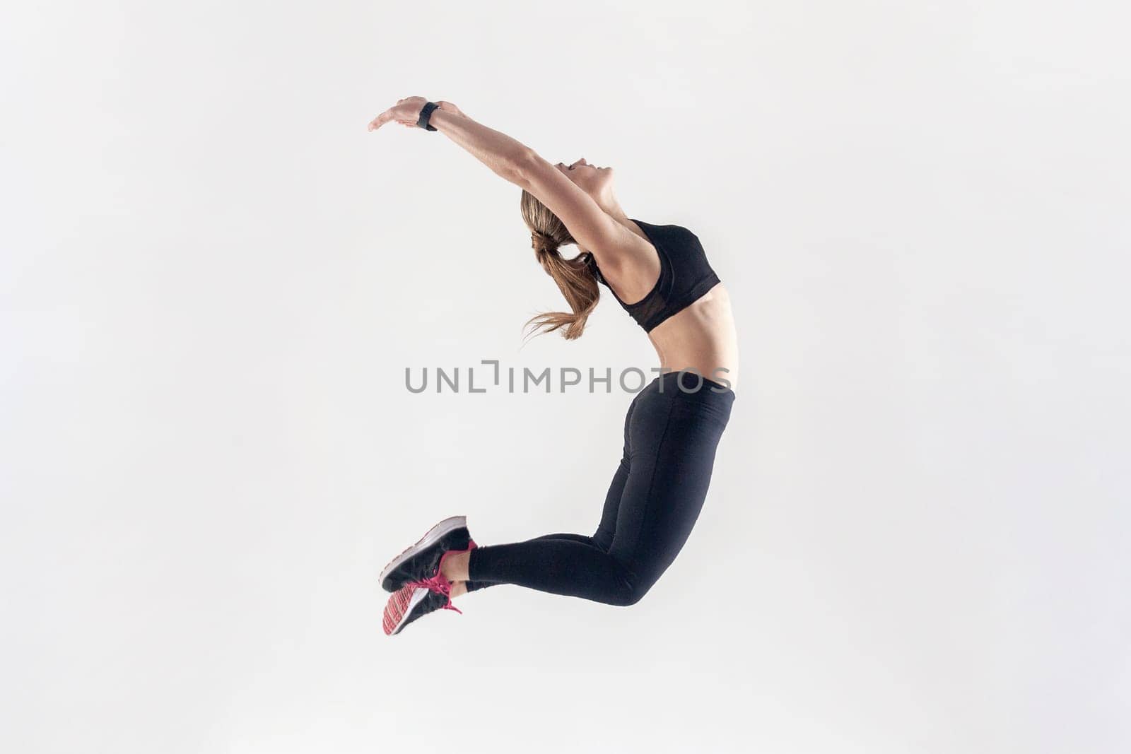 Side view portrait of athletic blonde woman with perfect body jumping high and keeping hands up, hands up, wearing black fitness clothing. Indoor studio shot isolated on gray background.