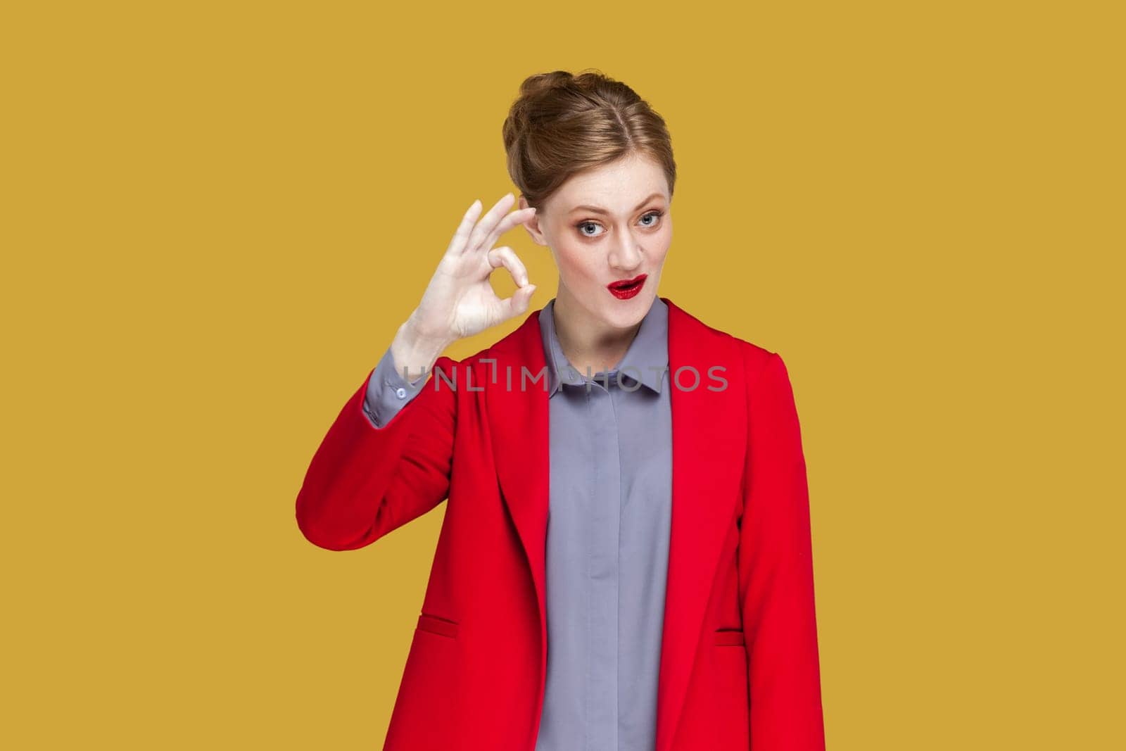 Okay, its fine. Woman says no problem, shows excellent gesture, has tender smile, gives recommendation, demonstrates positive approval, wearing jacket. Indoor studio shot isolated on yellow background