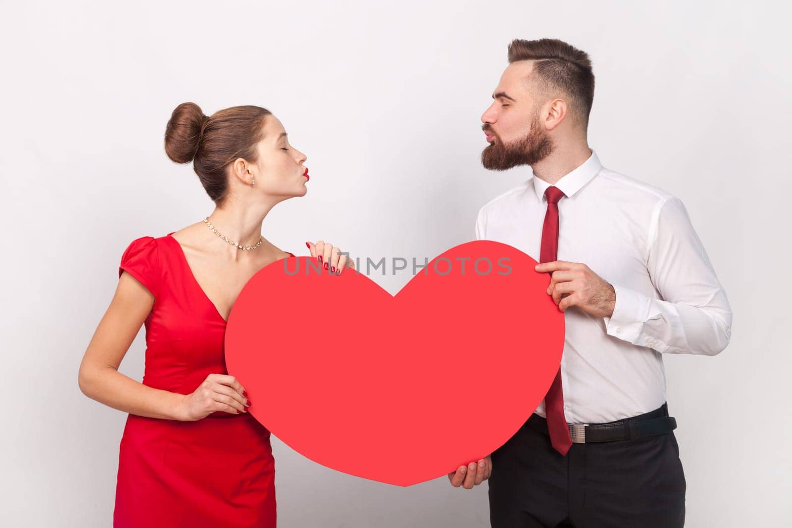 Portrait of lovely man in shirt and woman in red dress standing together, sending air kisses to each other, holding big heart. Indoor studio shot isolated on gray background.