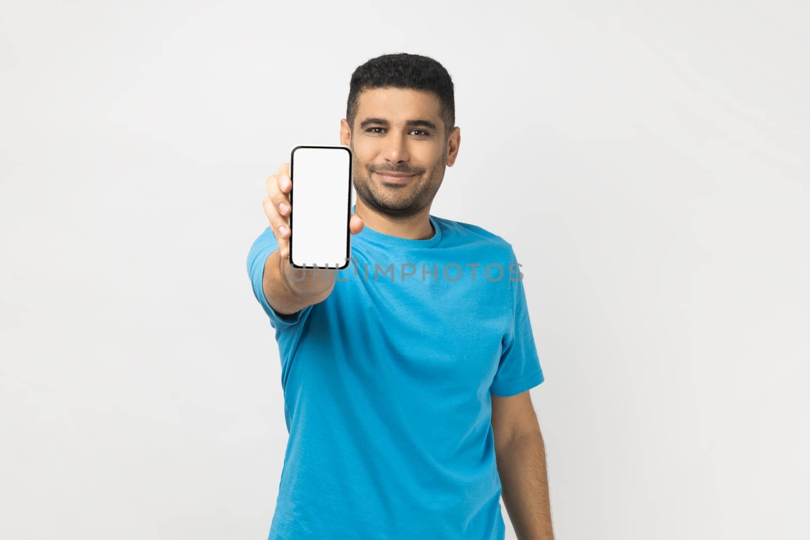 Portrait of cheerful satisfied unshaven man wearing blue T- shirt standing holding out smart phone with empty screen, copy space for advertisement. Indoor studio shot isolated on gray background.