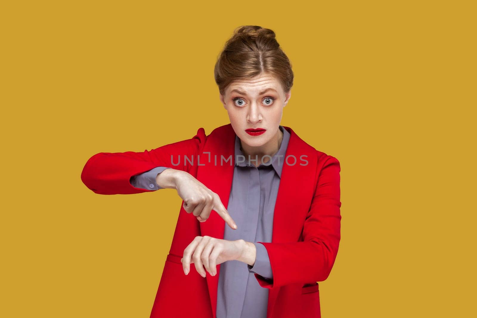 You are late. Portrait of serious strict woman with red lips standing pointing finger to her smartwatch, looking at camera, wearing red jacket. Indoor studio shot isolated on yellow background.