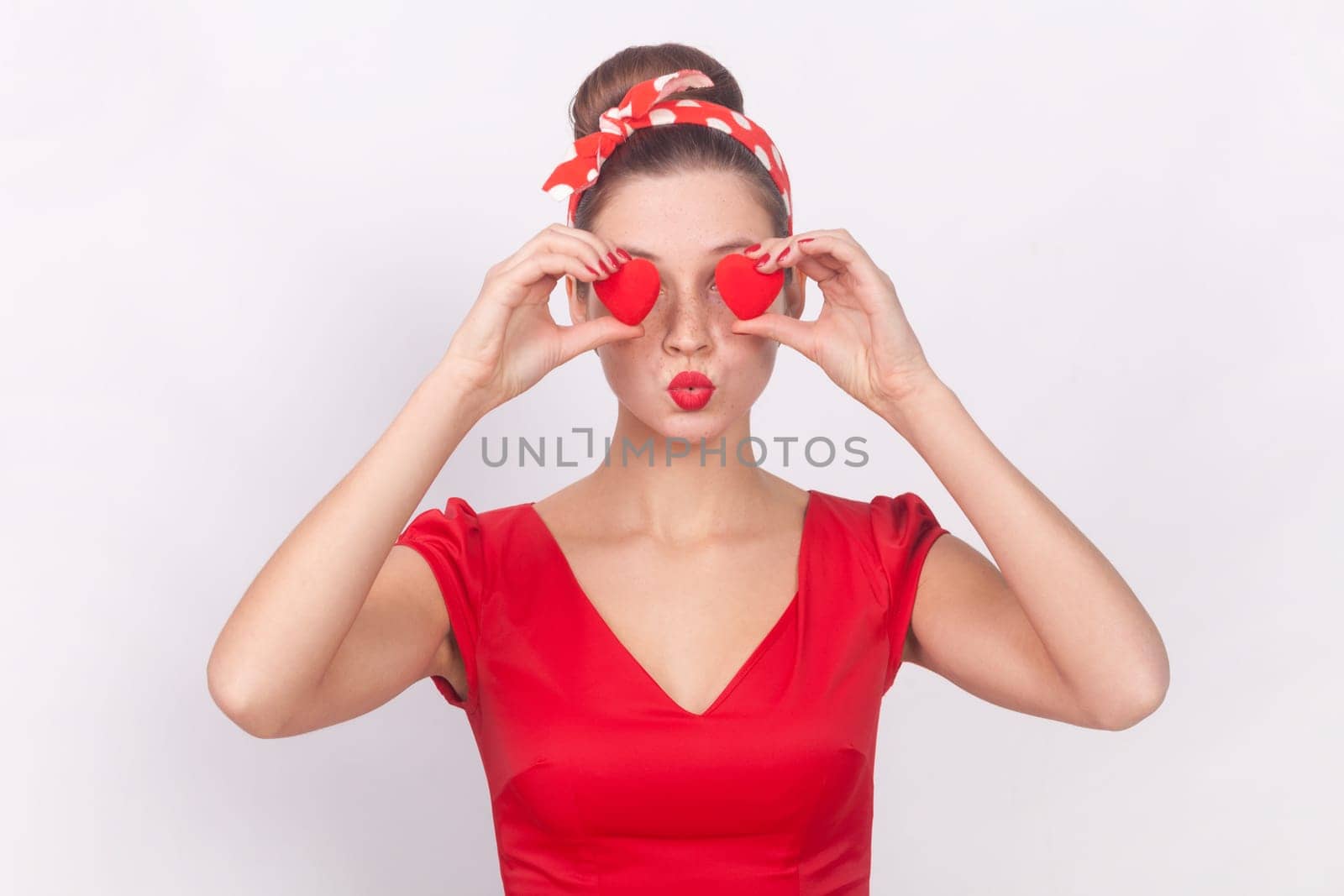 Portrait of romantic woman in red dress and head band covering her eyes with little red hearts, symbol of love, sending air kissing, keeps lips pout. Indoor studio shot isolated on gray background.