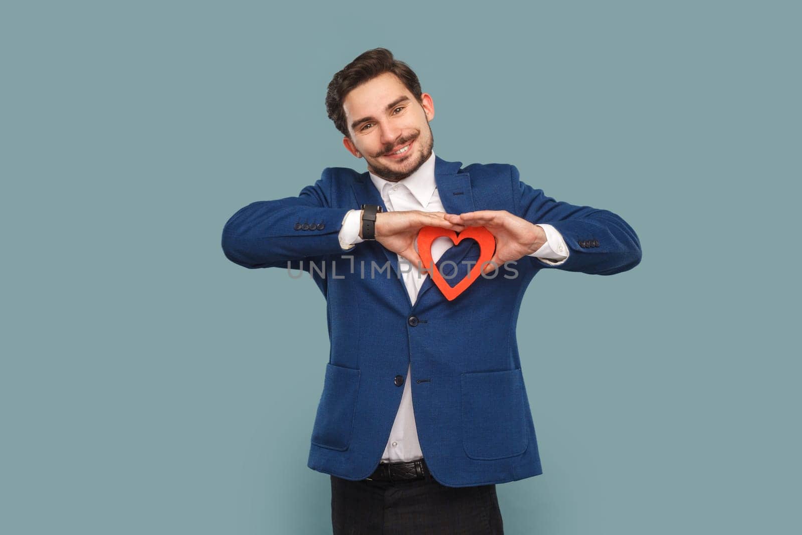 Romantic handsome man with mustache standing and showing red heart shaped figure, looking at camera with smile, wearing official style suit. Indoor studio shot isolated on light blue background.