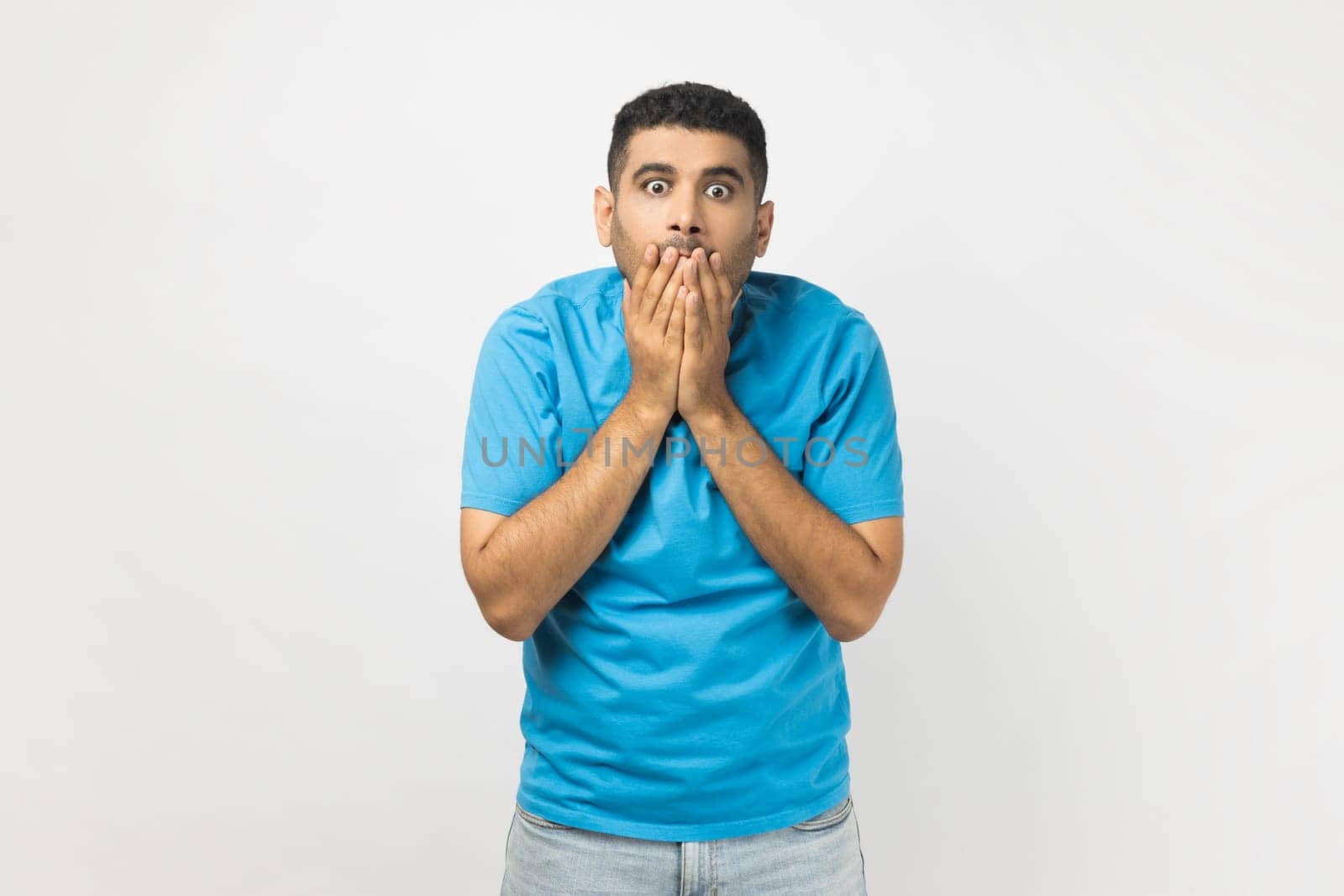 Portrait of astonished surprised man wearing blue T- shirt standing looking at camera with big eyes, covering mouth with palms, sees something shocked. Indoor studio shot isolated on gray background.