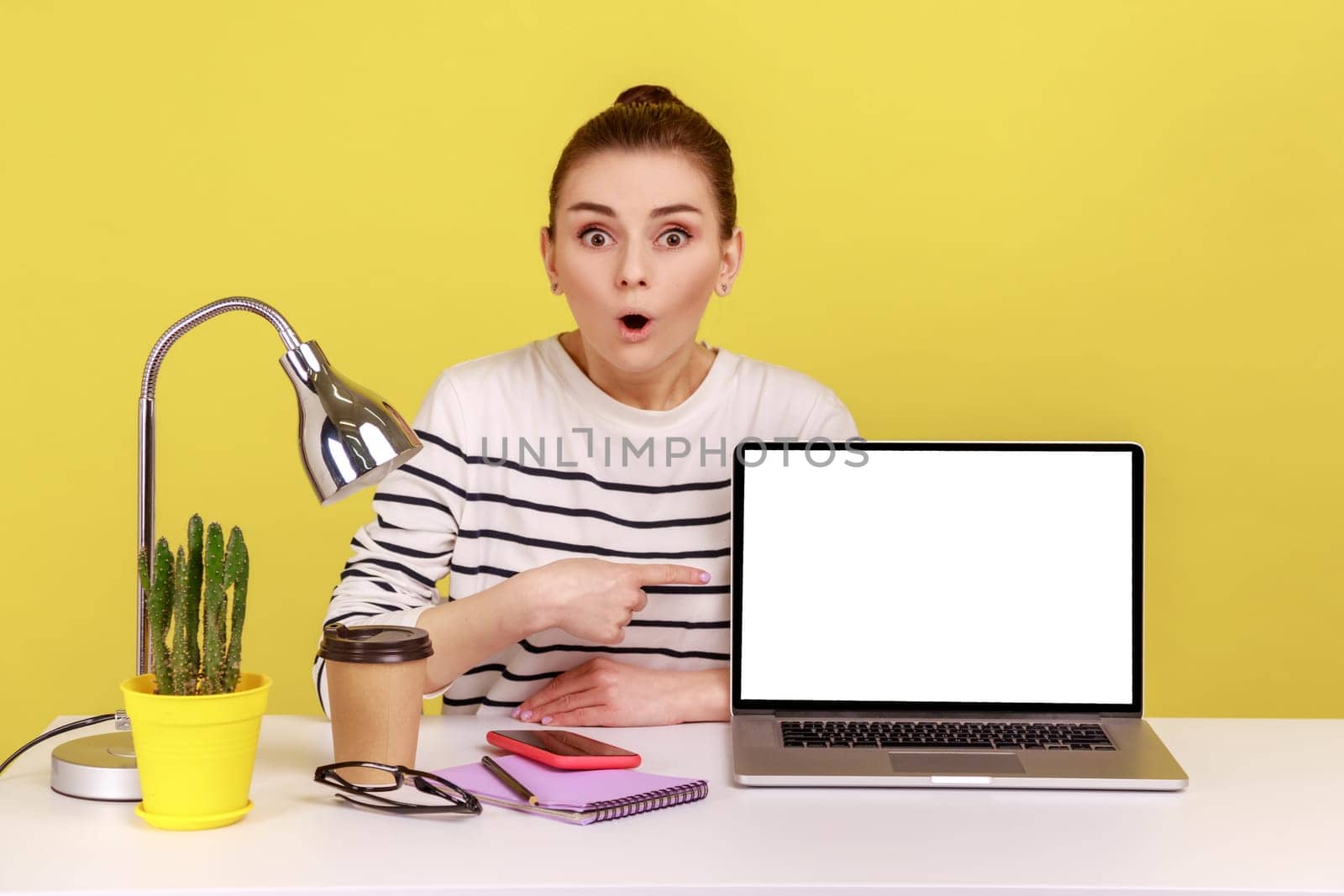 Shocked amazed young woman employee pointing empty laptop screen, mock up display for advertisement, online service, website promotion. Indoor studio studio shot isolated on yellow background.