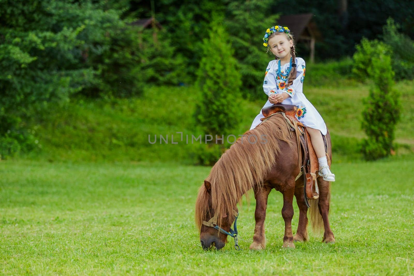A little girl in the Ukrainian national costume rides a pony on the lawn