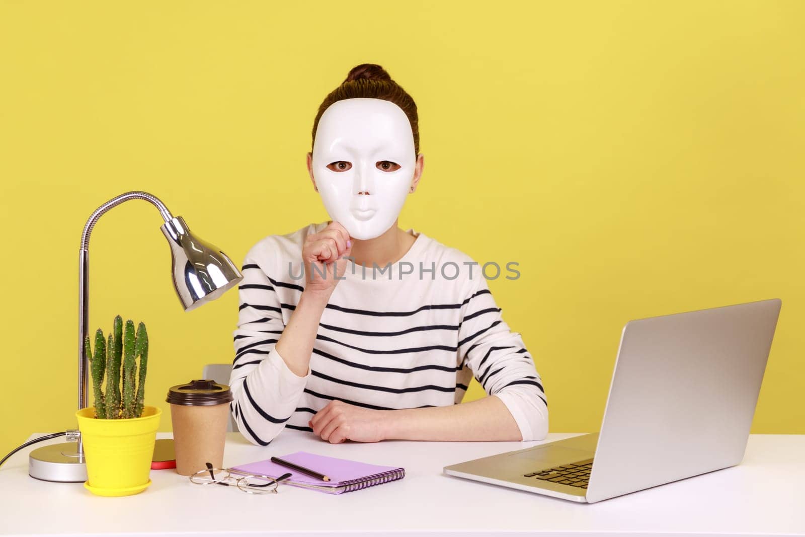 Portrait of unknown anonymous woman covering her face with white mask, hiding personality while sitting on workplace with laptop. Indoor studio studio shot isolated on yellow background.