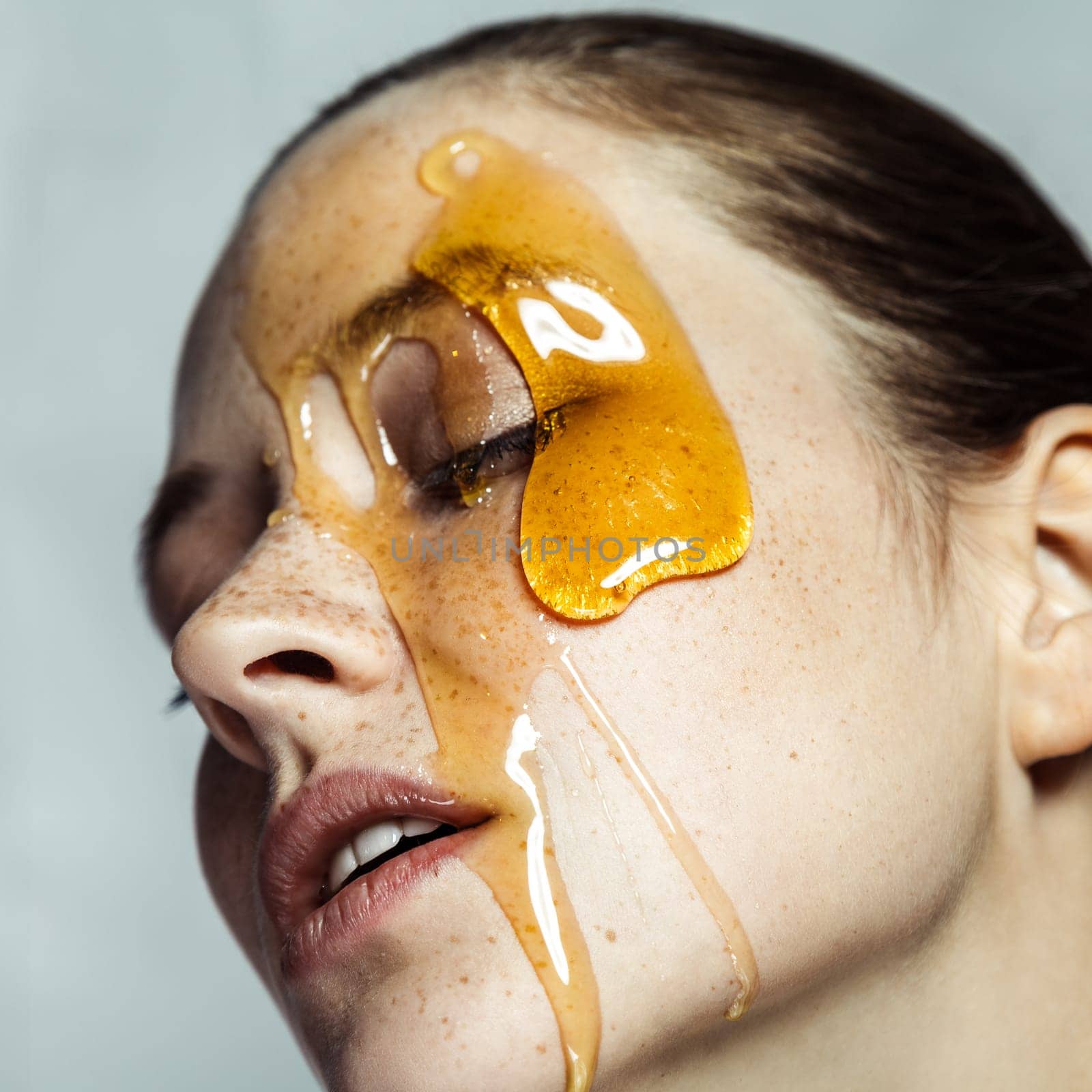 Closeup profile portrait of attractive young brunette woman with freckles, making honey homemade mask for skincare, posing with closed eyes. Indoor studio shot isolated on gray background.