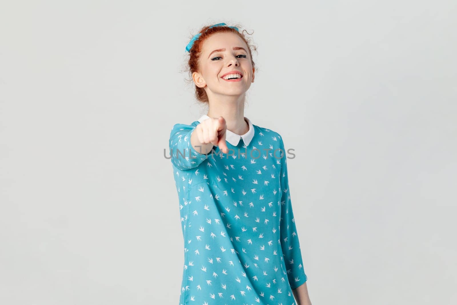Portrait of joyful cheerful red haired woman with bun hairstyle, standing indicating at camera, choosing you, laughing, wearing blue dress. Indoor studio shot isolated on gray background.