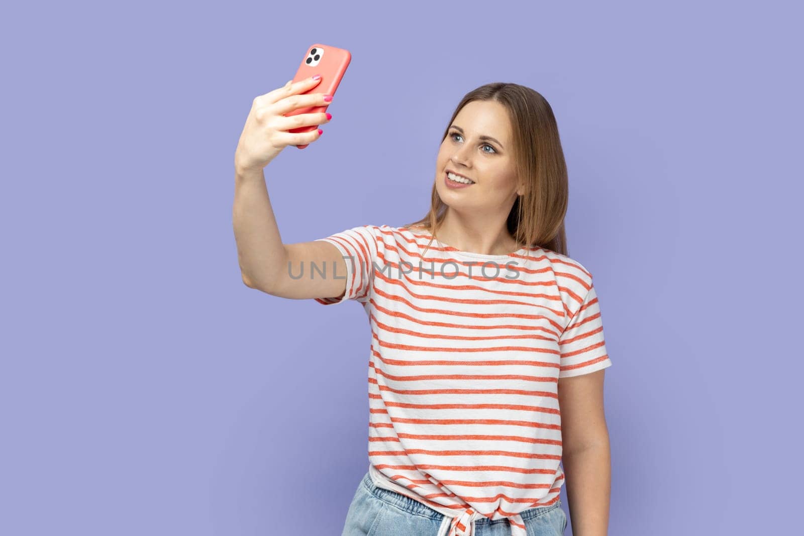 Portrait of smiling blond woman in striped T-shirt standing with phone and having video call or broadcasting livestream, looking smiling at screen. Indoor studio shot isolated on purple background.