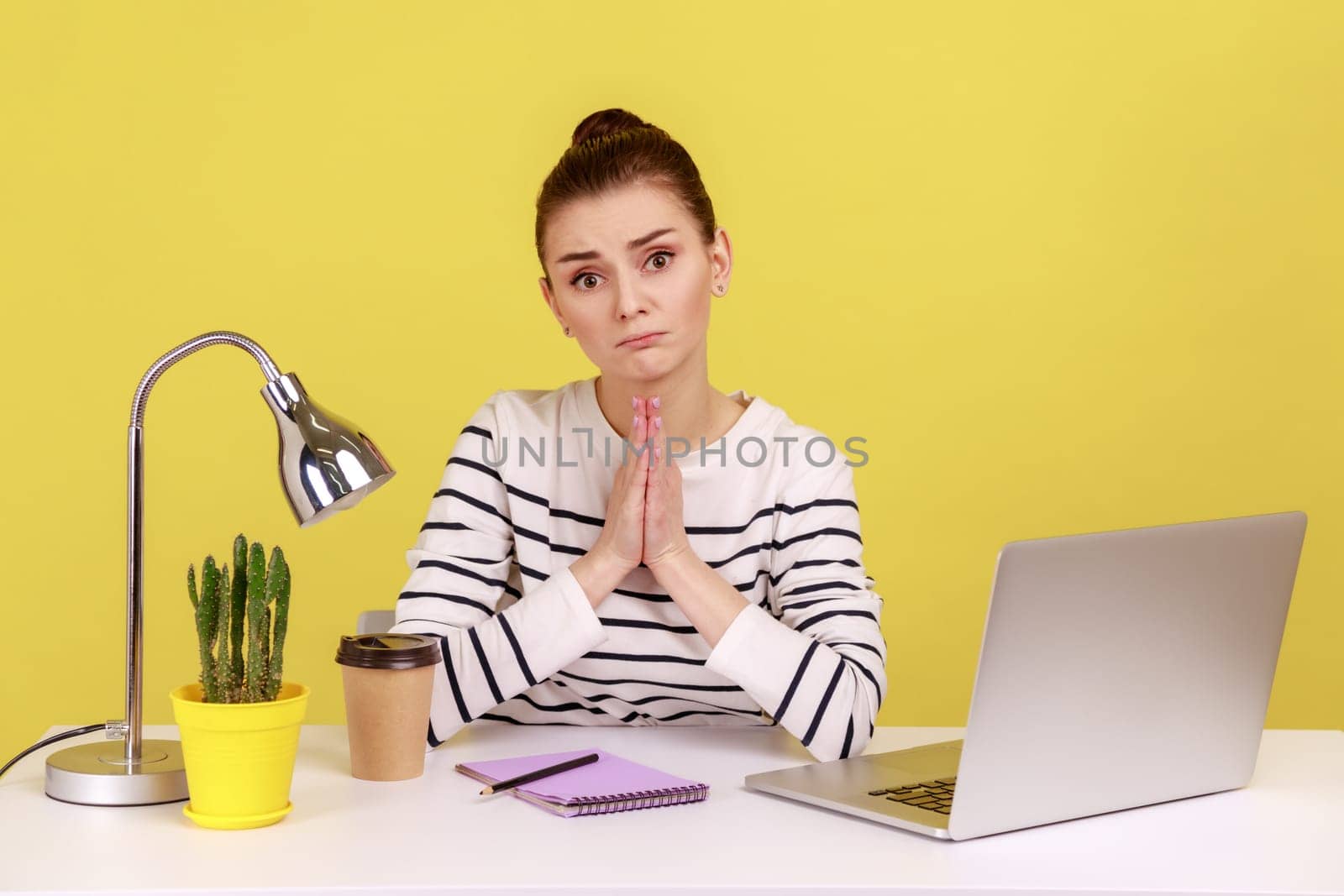Attractive woman manager sitting at workplace and holding hands in praying gesture, asking heartily, pleading with desperate face. Indoor studio studio shot isolated on yellow background.