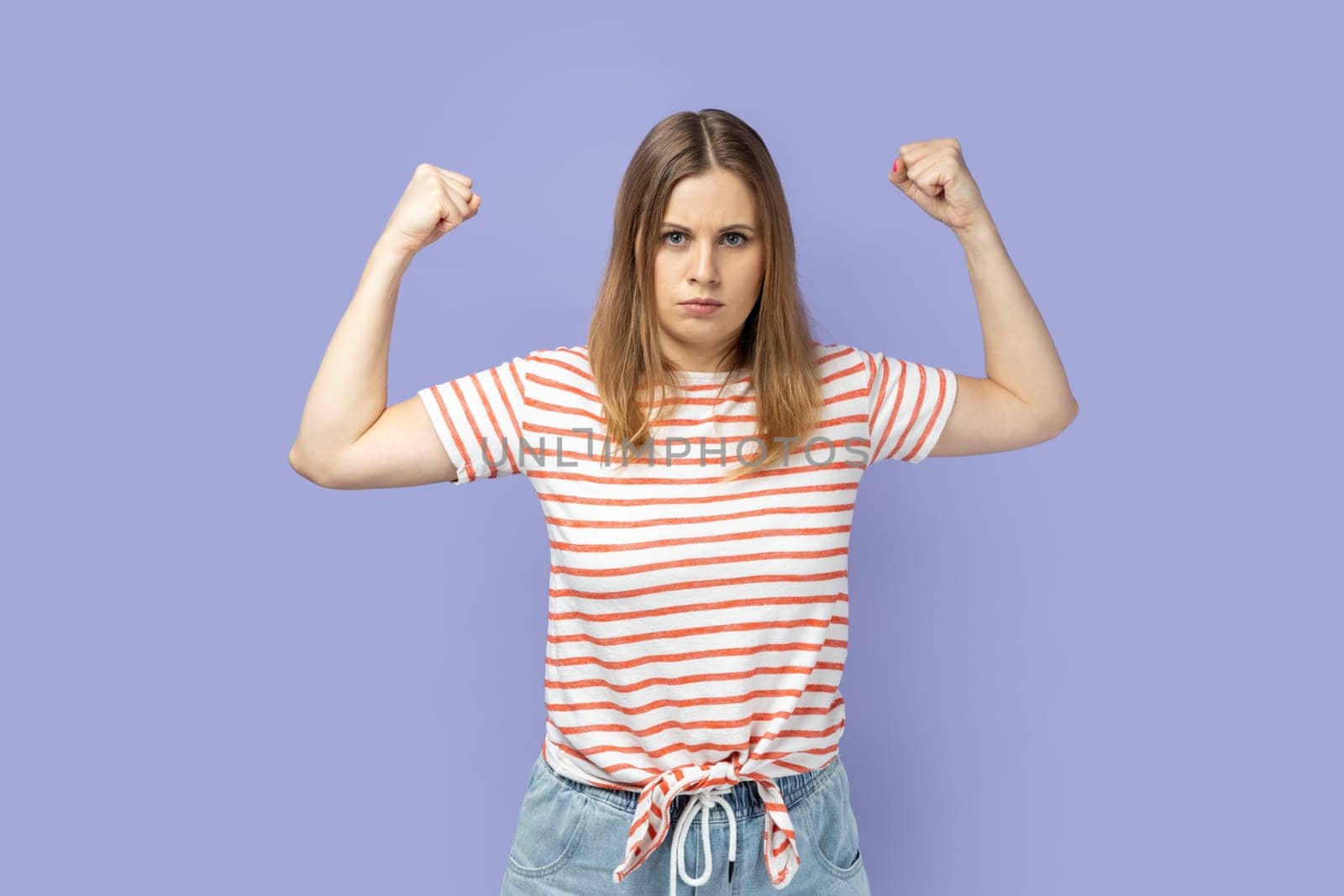 Portrait of strong confident blond woman wearing striped T-shirt standing with raised arms, showing her biceps and triceps, looking at camera. Indoor studio shot isolated on purple background.