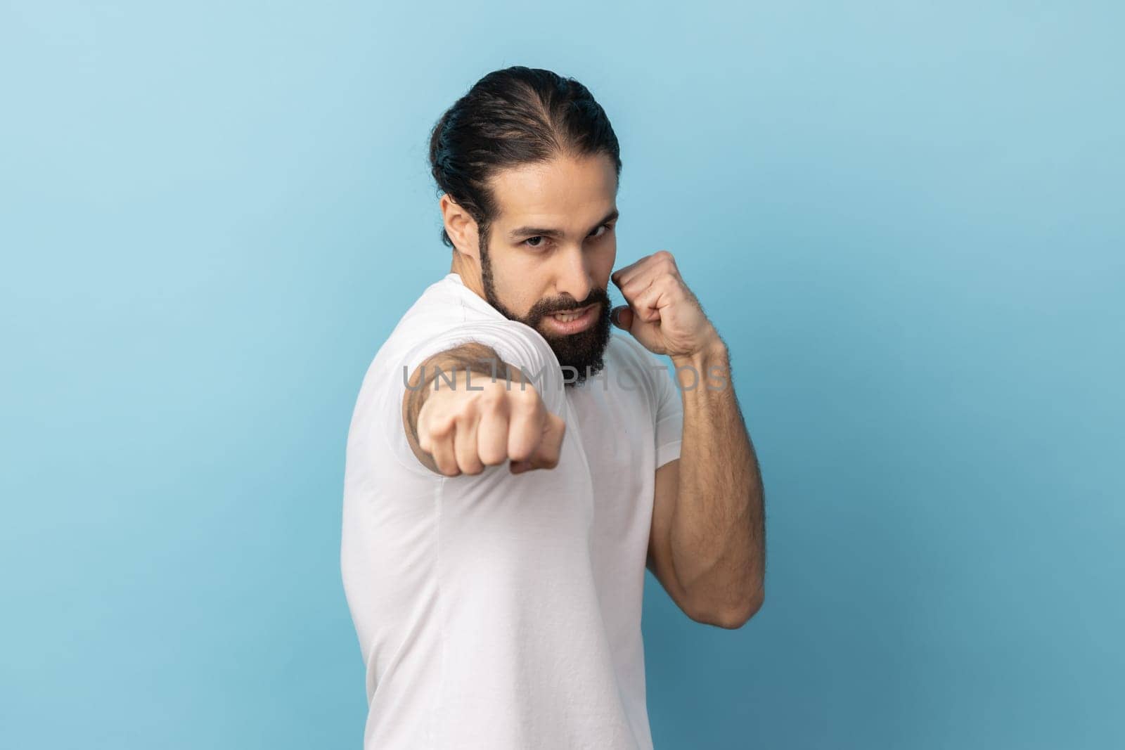 Portrait of angry aggressive man with beard wearing white T-shirt being ready to punch, boxing with clenched fists, fighting with somebody. Indoor studio shot isolated on blue background.