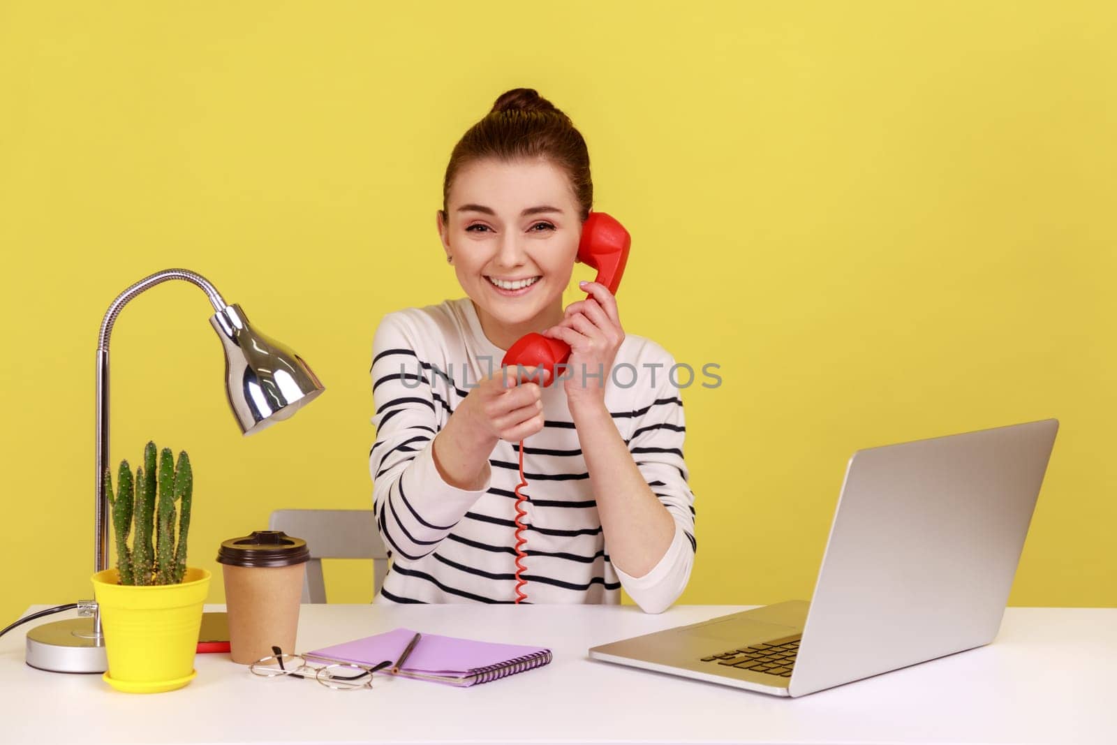 Woman secretary making calls on landline telephone working on laptop sitting at office, looking and pointing at camera with toothy smile. Indoor studio studio shot isolated on yellow background.