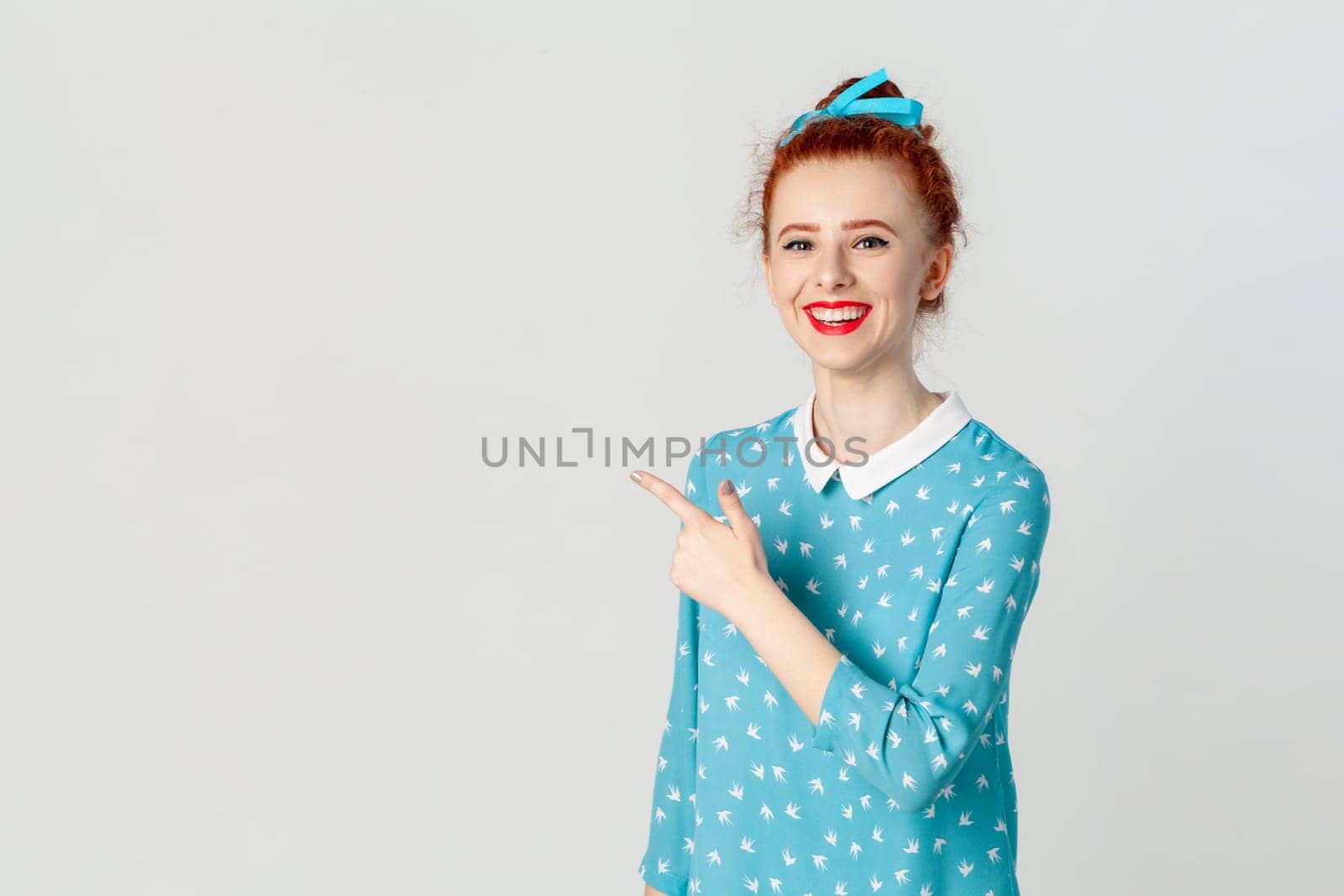 Portrait of smiling delighted ginger woman with bun hairstyle, pointing aside at advertisement area, copy space for promotion, wearing blue dress. Indoor studio shot isolated on gray background.