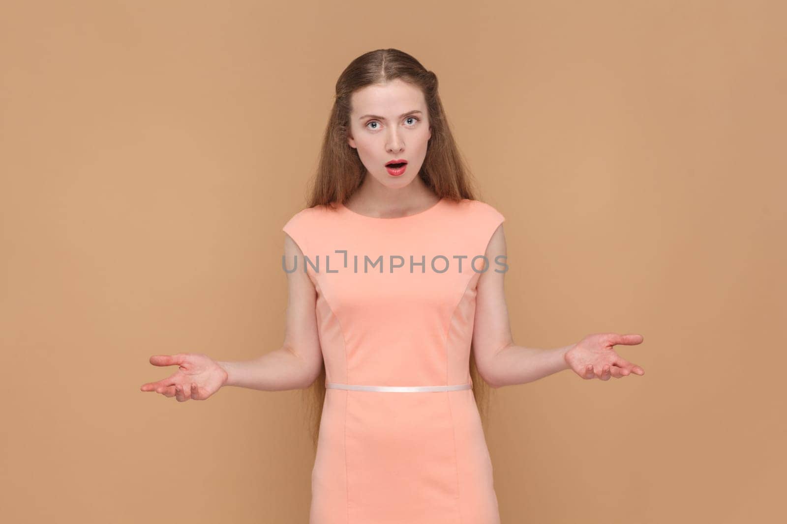 Portrait of angry shocked upset woman with long hair standing with raised arms, asking what, looking at camera, wearing elegant dress. Indoor studio shot isolated on brown background.