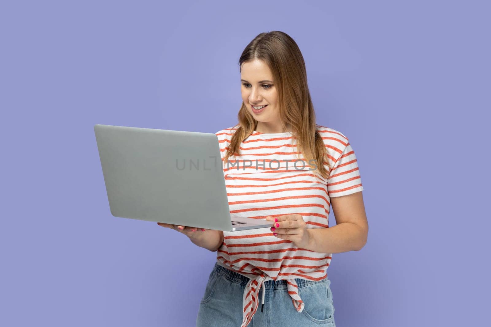 Portrait of smiling positive blond woman wearing striped T-shirt working on laptop, looking at display, enjoying her online work, reading information. Indoor studio shot isolated on purple background.