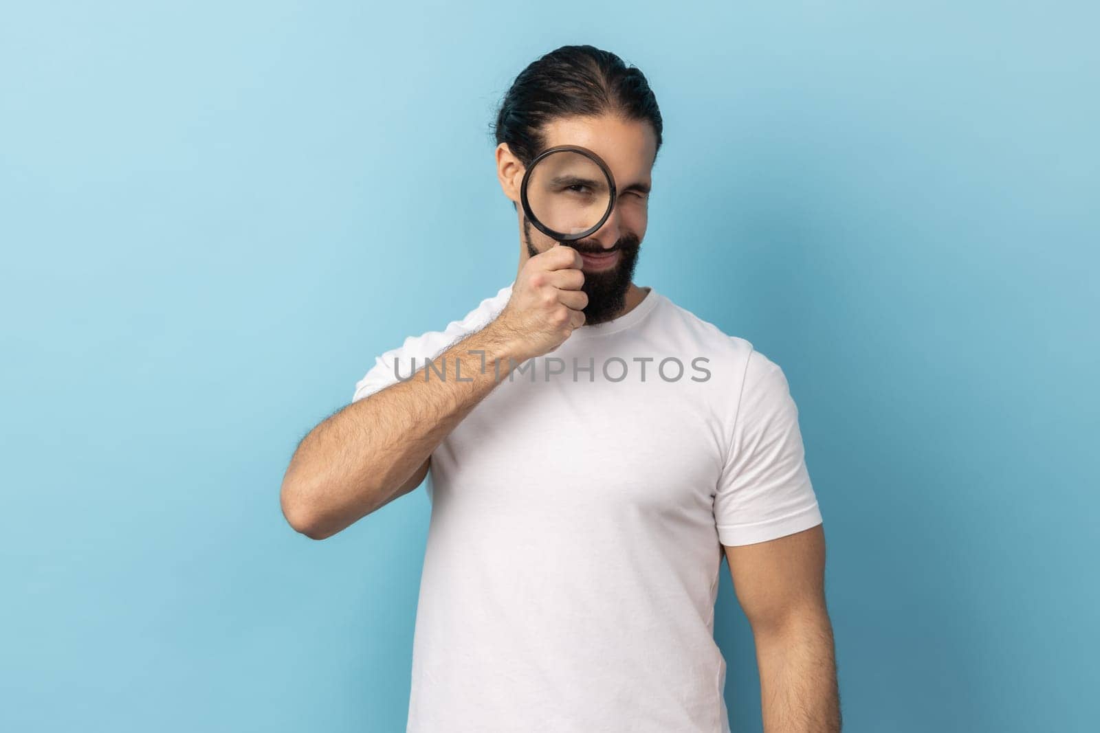 Portrait of man with beard wearing white T-shirt standing, holding magnifying glass and looking at camera with big zoom eye, verifying authenticity. Indoor studio shot isolated on blue background.