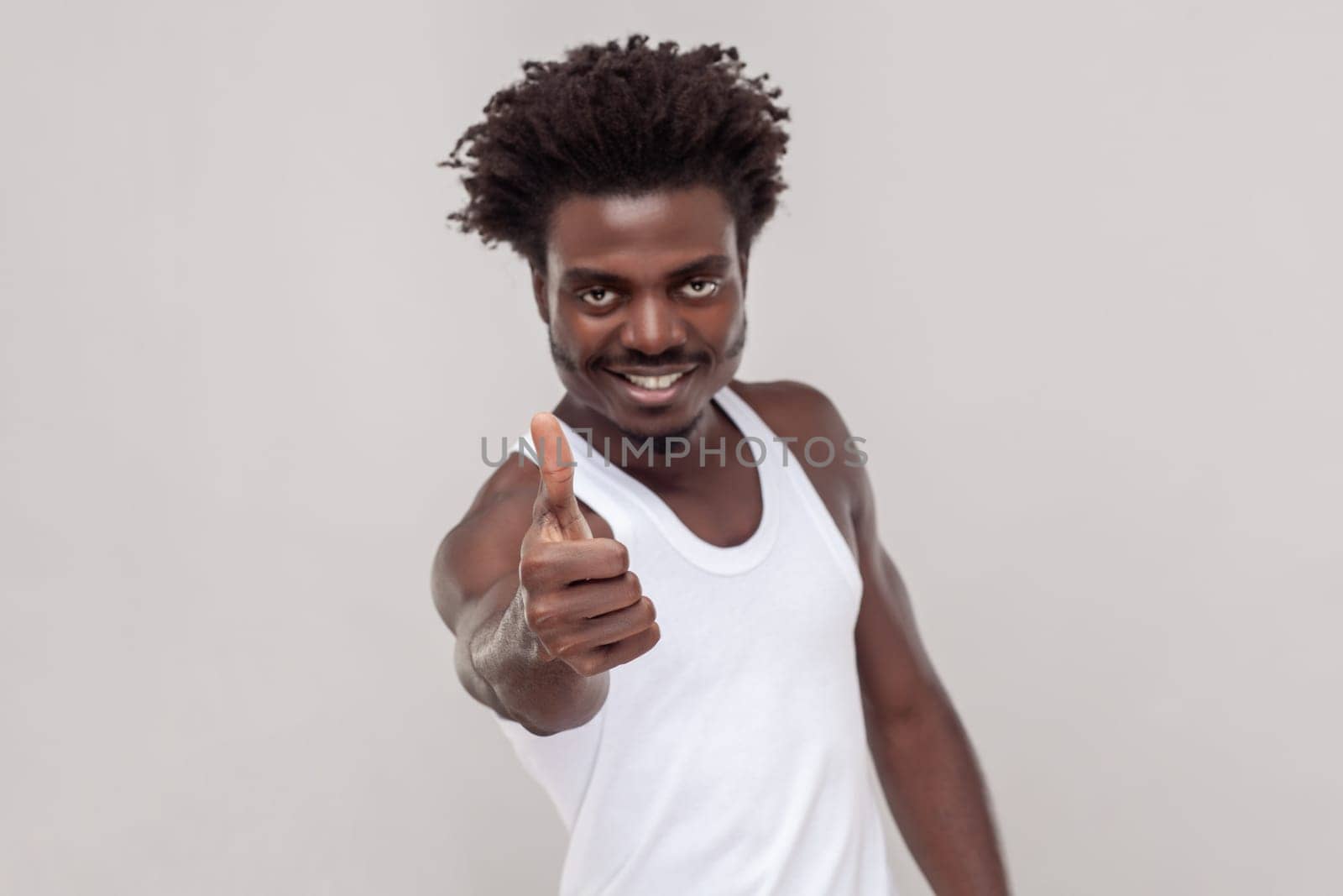 Very good. Pleased nice looking man with Afro hairstyle showing thumb up, gives positive feedback, demonstrates approval, wearing white T-shirt. Indoor studio shot isolated on gray background.